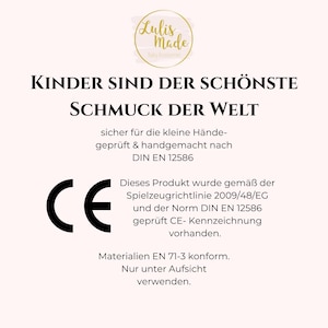 Op de afbeelding: Een witte achtergrond met zwarte tekst die "Kinder sind der sch&ouml;nste Schmuck der Welt" luidt. Onder de tekst bevindt zich een zwart-wit CE-teken en meer tekst in het Duits over veiligheidsnormen en materialen.
