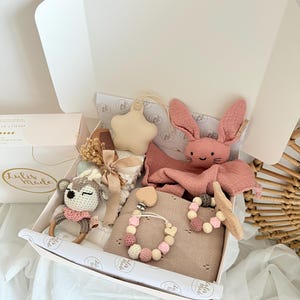 May include: A gift box filled with baby items, including a pink bunny plush, a star-shaped ornament, a crochet deer rattle, a blanket, and a pacifier clip. The box is white with a pink interior and is lined with patterned paper.
