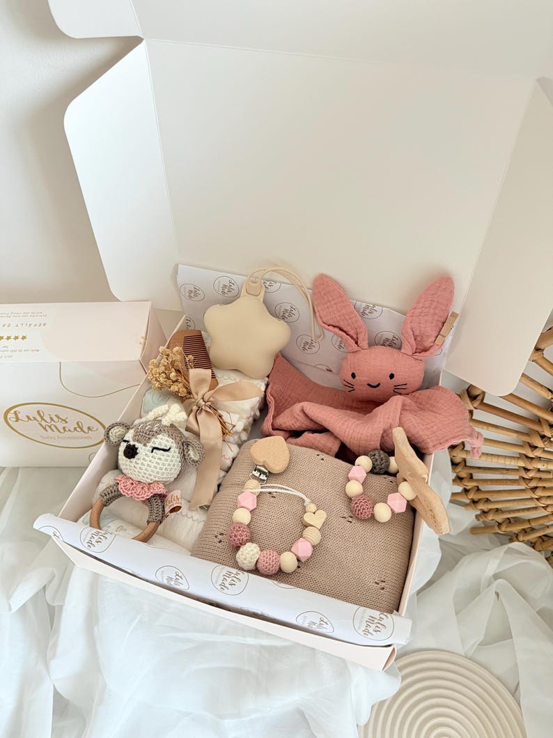 May include: A gift box filled with baby items, including a pink bunny plush, a star-shaped ornament, a crochet deer rattle, a blanket, and a pacifier clip. The box is white with a pink interior and is lined with patterned paper.