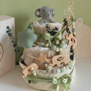 May include: A three-tiered baby diaper cake decorated with green ribbons, wooden elephant and star accents, and a crocheted gray elephant topper. The cake is adorned with eucalyptus sprigs and a string of wooden beads.