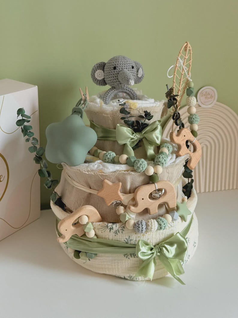 May include: A three-tiered baby diaper cake decorated with green ribbons, wooden elephant and star accents, and a crocheted gray elephant topper. The cake is adorned with eucalyptus sprigs and a string of wooden beads.