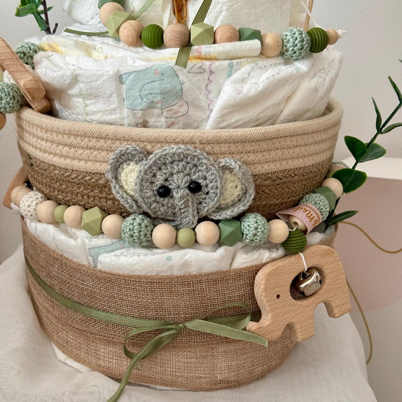 Op de afbeelding: Een luiercake versierd met groene en bruine kralen, een gehaakte olifant en een houten olifantenrammelaar. De cake is ingepakt in jute en vastgebonden met een groen lint.
