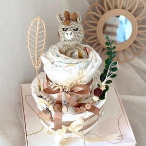 Peut inclure: Un g&acirc;teau &agrave; couches &agrave; deux &eacute;tages d&eacute;cor&eacute; d'un jouet girafe en crochet, de perles en bois et d'un ruban brun. Le g&acirc;teau est dans une bo&icirc;te blanche avec le texte "Hello Baby" et un c&oelig;ur.