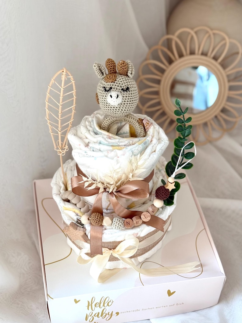Peut inclure: Un g&acirc;teau &agrave; couches &agrave; deux &eacute;tages d&eacute;cor&eacute; d'un jouet girafe en crochet, de perles en bois et d'un ruban brun. Le g&acirc;teau est dans une bo&icirc;te blanche avec le texte "Hello Baby" et un c&oelig;ur.