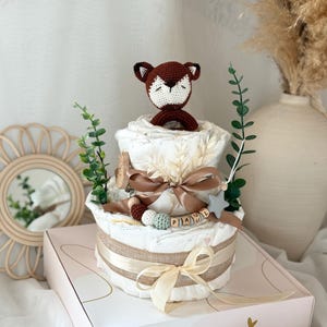Peut inclure: Un g&acirc;teau &agrave; couches &agrave; deux &eacute;tages d&eacute;cor&eacute; d'un jouet en crochet en forme de renard, d'une &eacute;toile et d'une &eacute;tiquette de nom en bois qui dit "PAUL". Le g&acirc;teau est envelopp&eacute; d'un ruban en toile de jute et d'un ruban cr&egrave;me. Le g&acirc;teau est pos&eacute; sur une bo&icirc;te blanche avec le texte "Hello Baby" et "Zuckers&uuml;sse Sachen warten auf Dich".