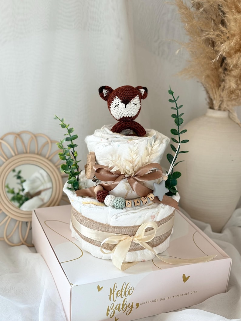 Peut inclure: Un g&acirc;teau &agrave; couches &agrave; deux &eacute;tages d&eacute;cor&eacute; d'un jouet en crochet en forme de renard, d'une &eacute;toile et d'une &eacute;tiquette de nom en bois qui dit "PAUL". Le g&acirc;teau est envelopp&eacute; d'un ruban en toile de jute et d'un ruban cr&egrave;me. Le g&acirc;teau est pos&eacute; sur une bo&icirc;te blanche avec le texte "Hello Baby" et "Zuckers&uuml;sse Sachen warten auf Dich".