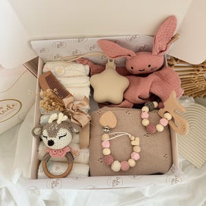 May include: A baby gift box filled with various items. Includes a pink plush bunny, a star-shaped rattle, a wooden comb, a crochet deer rattle, a pacifier clip, and a blanket. The box is white with a patterned interior.