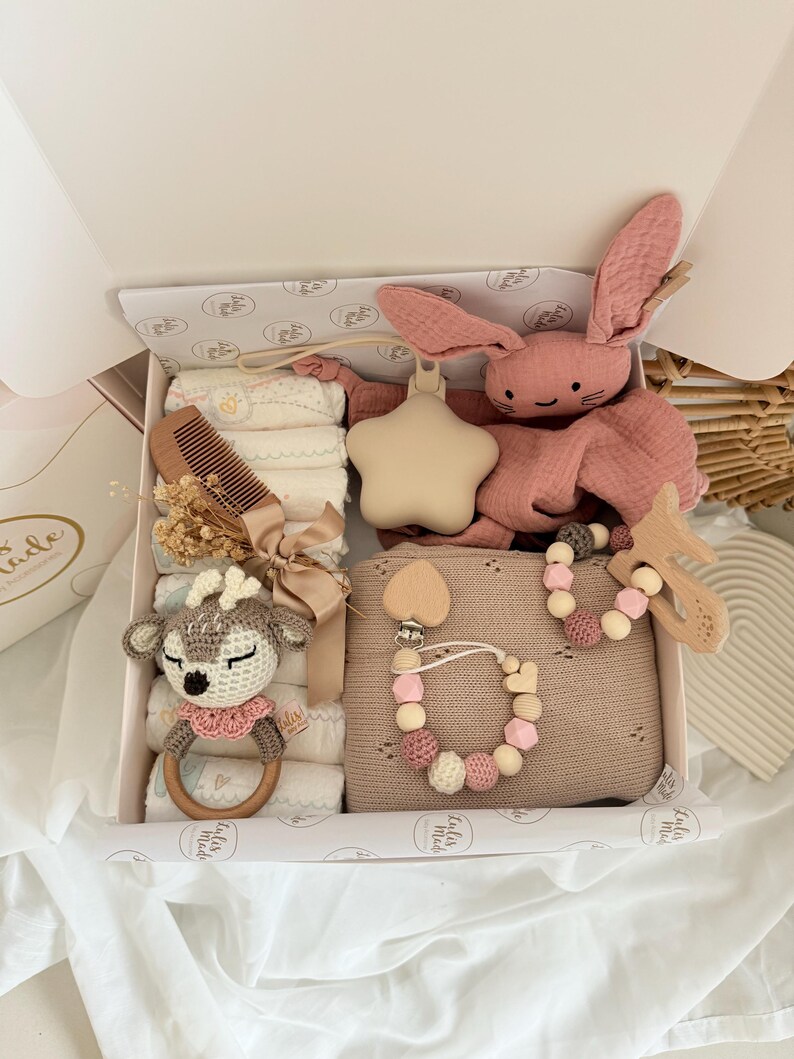 May include: A baby gift box filled with various items. Includes a pink plush bunny, a star-shaped rattle, a wooden comb, a crochet deer rattle, a pacifier clip, and a blanket. The box is white with a patterned interior.