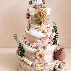 May include: A three-tiered diaper cake decorated with a crocheted deer, wooden beads, and a heart-shaped pendant. The cake includes a woven basket base, a bow, and a pair of baby booties. The cake is decorated with dried flowers and greenery.