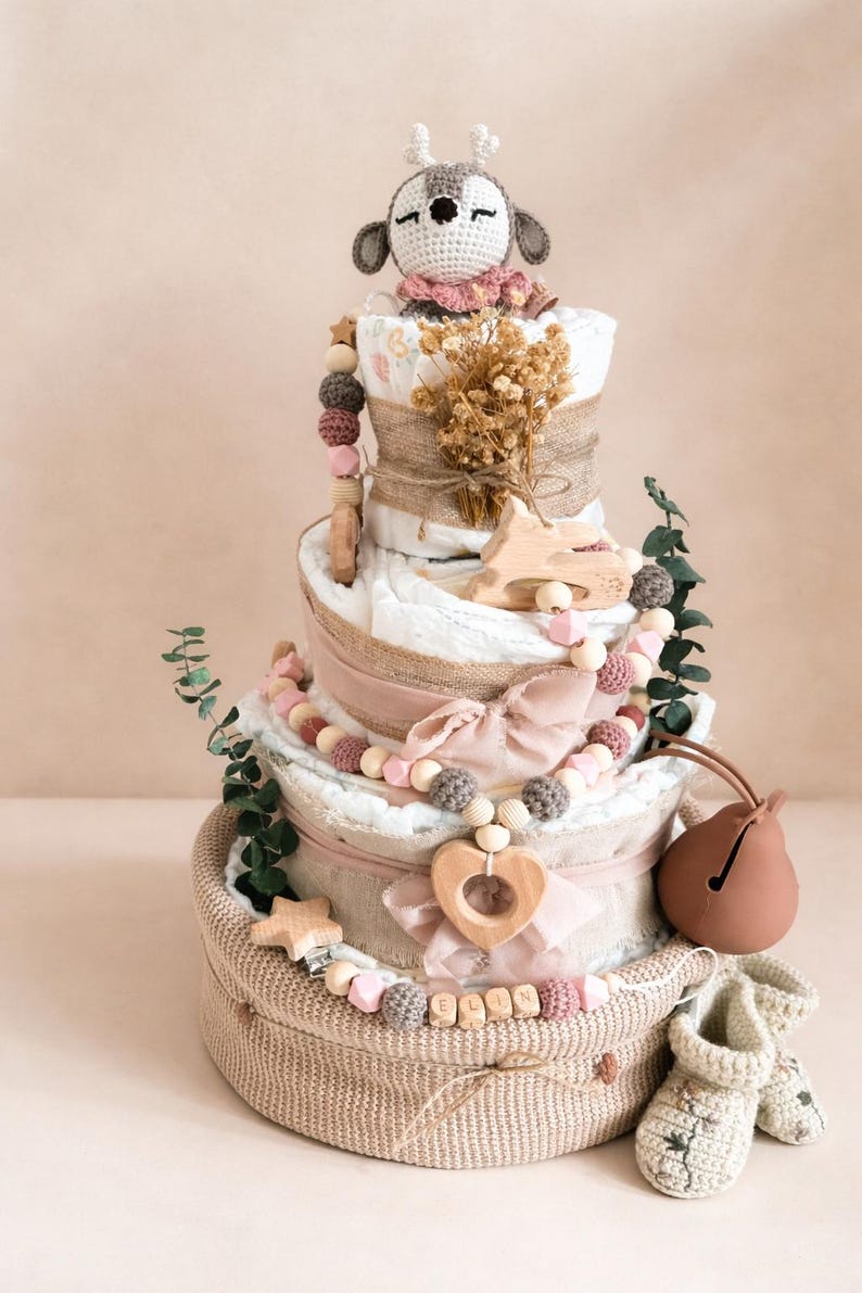 May include: A three-tiered diaper cake decorated with a crocheted deer, wooden beads, and a heart-shaped pendant. The cake includes a woven basket base, a bow, and a pair of baby booties. The cake is decorated with dried flowers and greenery.