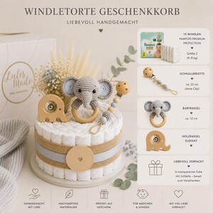 May include: A baby gift basket shaped like a cake, featuring a crochet elephant teether, wooden elephant rattle, and a pacifier clip. The cake is decorated with diapers, ribbons, and a wooden circle. The text on the box says "Lulis Made Baby Accessories".