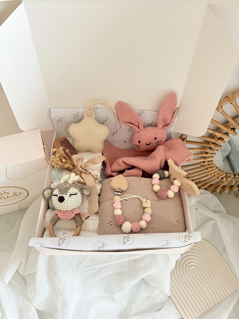 May include: A baby gift set in a white box, featuring a pink bunny plush, a star-shaped teether, a wooden deer rattle, a pacifier clip, and a blanket. The set includes a wooden comb and other baby essentials. The items are in shades of pink, beige, and white.