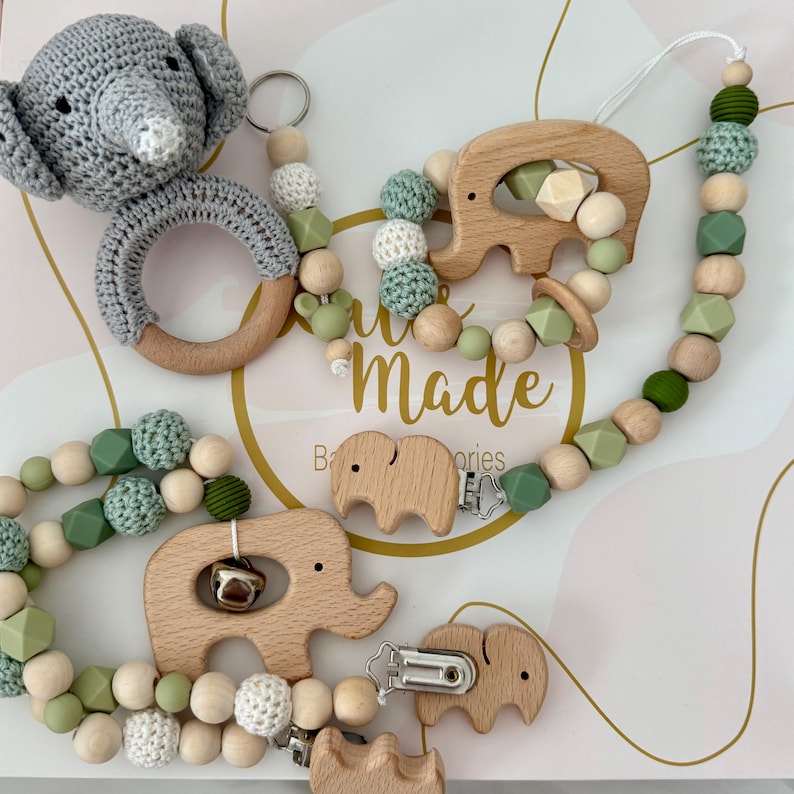 Op de afbeelding: Een groene en beige baby-bijtring met houten olifantvormen en kralen. De bijtring heeft een clip om aan kleding te bevestigen. De bijtring is handgemaakt en heeft de tekst "Hand Made" op de doos.