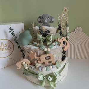 May include: A three-tiered baby gift set with a gray crocheted elephant topper. The set includes wooden elephant and star-shaped toys, green bows, and decorative beads. The base is wrapped in a floral patterned fabric.