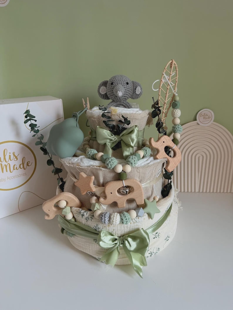 May include: A three-tiered baby gift set with a gray crocheted elephant topper. The set includes wooden elephant and star-shaped toys, green bows, and decorative beads. The base is wrapped in a floral patterned fabric.