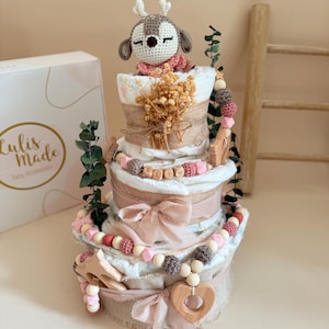 May include: A diaper cake with a crochet deer topper, burlap accents, and pink ribbons. The cake is decorated with wooden beads, a wooden heart, and the name "ELLIN" spelled out in wooden letters. A box with the text "Lulis Made Baby Accessories" is visible.