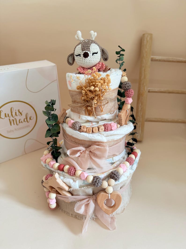 May include: A diaper cake with a crochet deer topper, burlap accents, and pink ribbons. The cake is decorated with wooden beads, a wooden heart, and the name "ELLIN" spelled out in wooden letters. A box with the text "Lulis Made Baby Accessories" is visible.