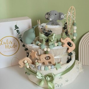 May include: A three-tiered baby diaper cake with a gray crocheted elephant topper. The cake is decorated with wooden animal toys, green bows, and a garland of beads. A box with the text "Lulis Made Baby Accessories" is on the left.
