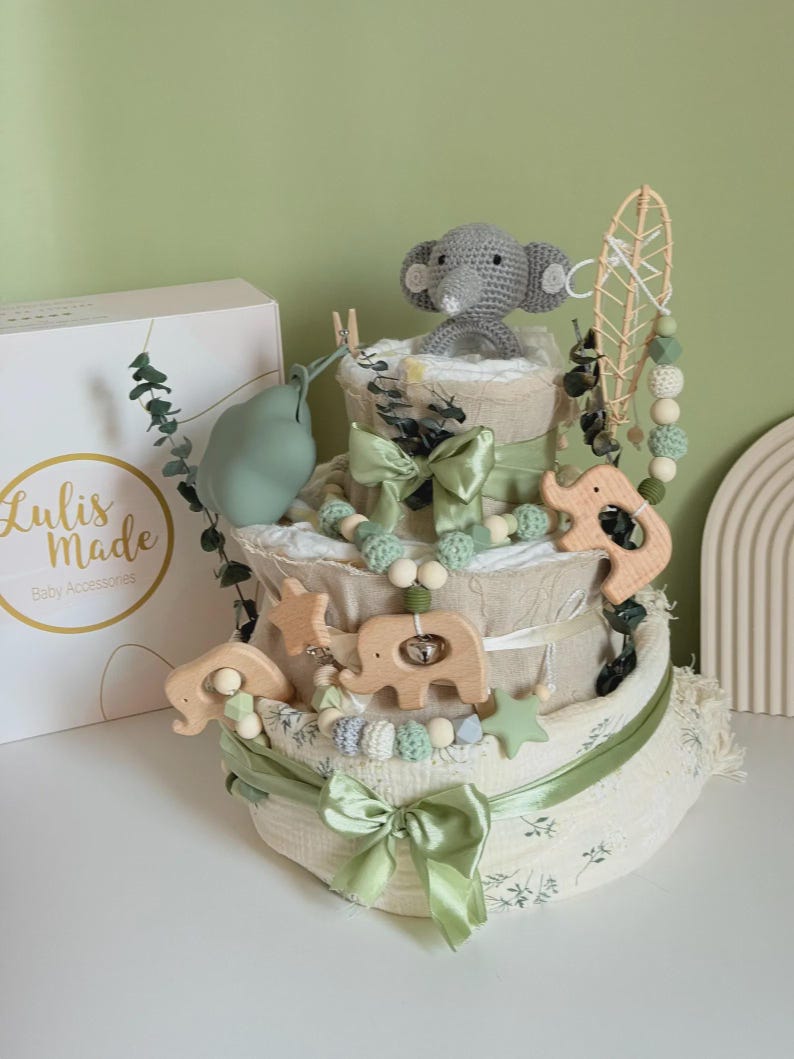 May include: A three-tiered baby diaper cake with a gray crocheted elephant topper. The cake is decorated with wooden animal toys, green bows, and a garland of beads. A box with the text "Lulis Made Baby Accessories" is on the left.