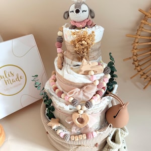 May include: A multi-tiered baby gift basket with a crochet deer topper, wooden beads, and a bell. The basket includes diapers, decorative ribbons, and small wooden accents. The color palette is soft, featuring beige, pink, and white.