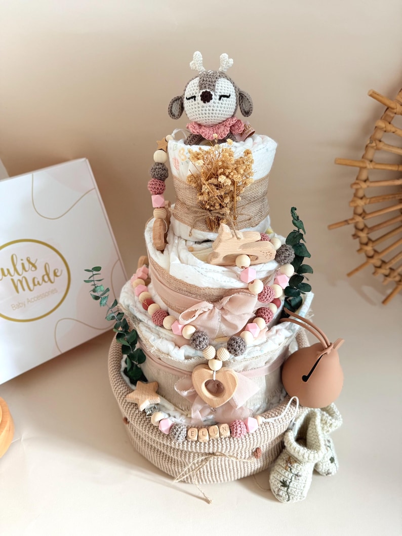 May include: A multi-tiered baby gift basket with a crochet deer topper, wooden beads, and a bell. The basket includes diapers, decorative ribbons, and small wooden accents. The color palette is soft, featuring beige, pink, and white.
