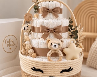 May include: A three-tier diaper cake in a woven basket with a face design. It features brown satin ribbons, a crocheted bear rattle, a star-shaped teether, and a wooden pacifier clip engraved with the name Clio, accented with dried floral stems.