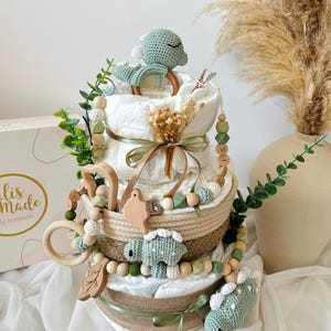May include: A three-tiered diaper cake decorated with green and beige accents. The cake features a crochet dinosaur, wooden beads, and a wooden ring. The cake is adorned with greenery and a ribbon. The cake is a baby shower gift.