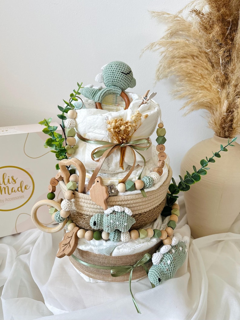 May include: A three-tiered diaper cake decorated with green and beige accents. The cake features a crochet dinosaur, wooden beads, and a wooden ring. The cake is adorned with greenery and a ribbon. The cake is a baby shower gift.