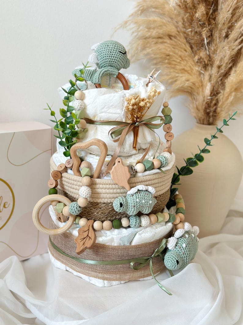 May include: A three-tiered diaper cake decorated with green and beige accents. The cake features crocheted dinosaur figures, wooden beads, and a variety of decorative elements, including a wooden leaf and a wooden ring. The cake is wrapped in burlap and ribbon.