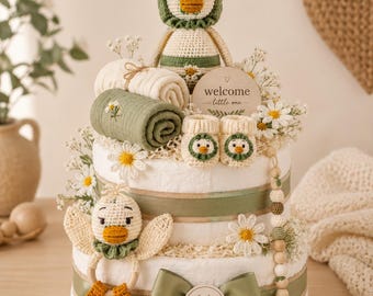 May include: A two-tier diaper cake decorated with sage green ribbons, a Lulis Made tag, and a wooden welcome little one sign. It features crochet duck toys, rolled muslin blankets, baby booties, white daisy flowers, and a wooden bead pacifier clip.