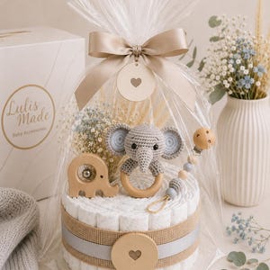 May include: A baby gift set featuring a diaper cake with a crocheted elephant rattle, wooden elephant toy, and a pacifier clip. The cake is wrapped in cellophane and tied with a beige ribbon. The gift set includes the text "Lulis Made Baby Accessories".