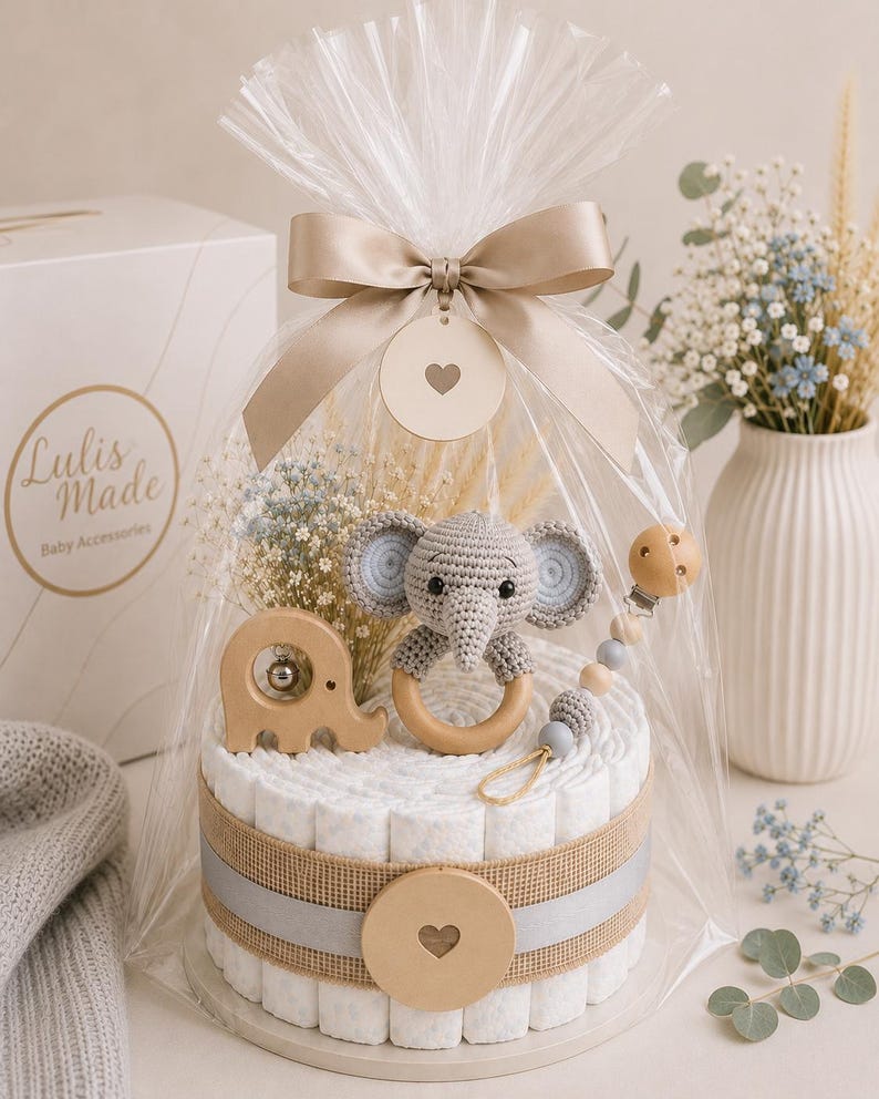 May include: A baby gift set featuring a diaper cake with a crocheted elephant rattle, wooden elephant toy, and a pacifier clip. The cake is wrapped in cellophane and tied with a beige ribbon. The gift set includes the text "Lulis Made Baby Accessories".