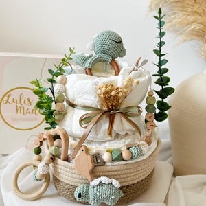 May include: A diaper cake with a woven basket base, adorned with a crocheted turtle, wooden beads, and greenery. The cake features layers of diapers, a wooden teether, and a ribbon. The box in the background has the text "Lulis Made Baby Accessories".