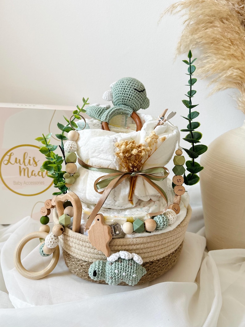 May include: A diaper cake with a woven basket base, adorned with a crocheted turtle, wooden beads, and greenery. The cake features layers of diapers, a wooden teether, and a ribbon. The box in the background has the text "Lulis Made Baby Accessories".