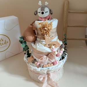 May include: A three-tiered diaper cake with a deer topper, burlap accents, and pink ribbons. The cake is decorated with wooden stars, beads, and greenery. A pear-shaped teether and a box with the text "is made Accessories" are also present.