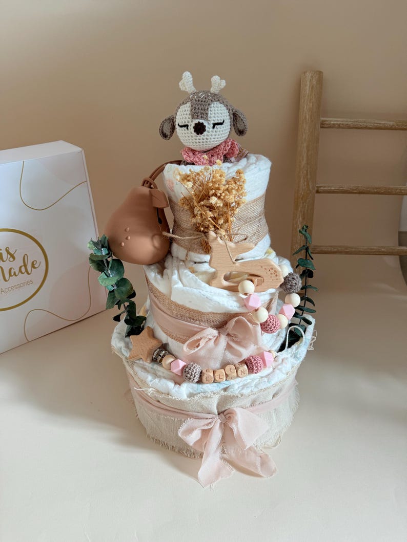 May include: A three-tiered diaper cake with a deer topper, burlap accents, and pink ribbons. The cake is decorated with wooden stars, beads, and greenery. A pear-shaped teether and a box with the text "is made Accessories" are also present.