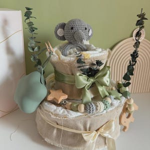 May include: A two-tiered diaper cake decorated with a gray crocheted elephant, green ribbons, and wooden embellishments. The cake is wrapped in burlap and adorned with eucalyptus branches and a green pacifier.