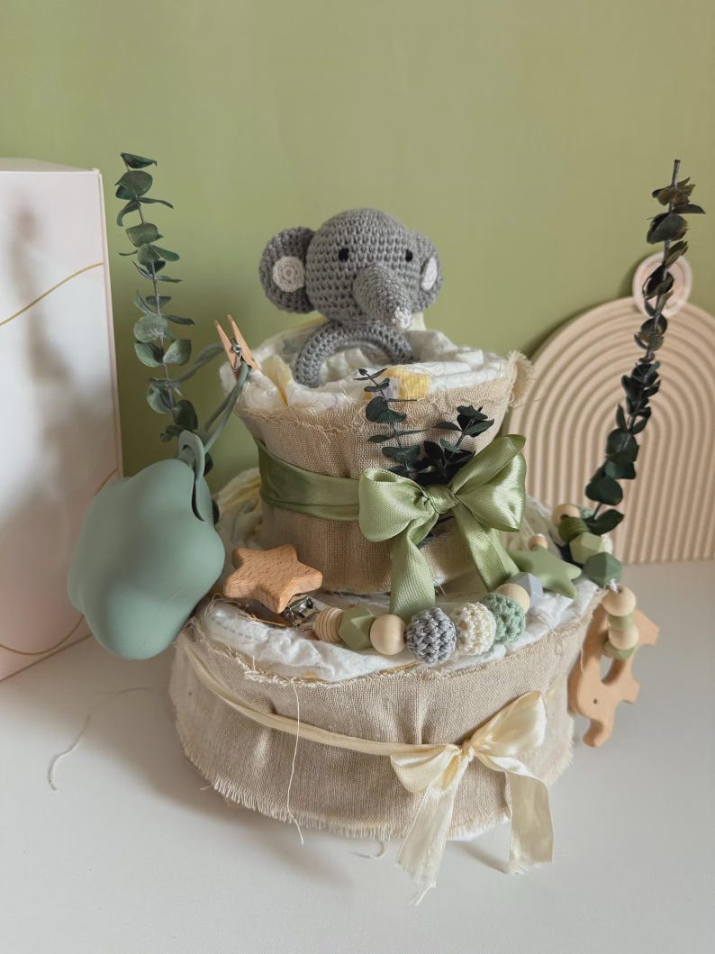May include: A two-tiered diaper cake decorated with a gray crocheted elephant, green ribbons, and wooden embellishments. The cake is wrapped in burlap and adorned with eucalyptus branches and a green pacifier.