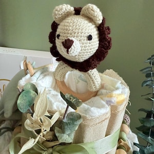 May include: A handmade baby shower gift featuring a crochet lion topper. The diaper cake is wrapped in burlap and adorned with green ribbon, wooden beads, and faux greenery. The lion has a tan face and brown mane.