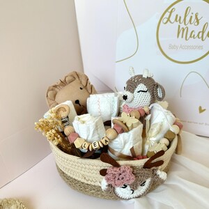 May include: A wicker basket filled with baby items, including a crocheted deer toy, a lion plush, a wooden name tag, and a teething toy. The basket is decorated with a crocheted deer face.