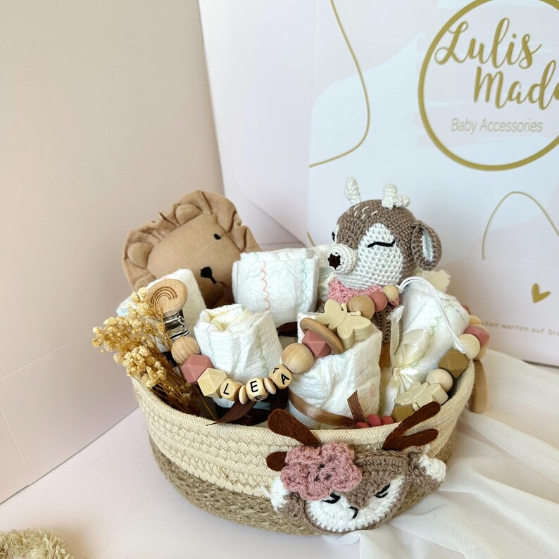 May include: A wicker basket filled with baby items, including a crocheted deer toy, a lion plush, a wooden name tag, and a teething toy. The basket is decorated with a crocheted deer face.