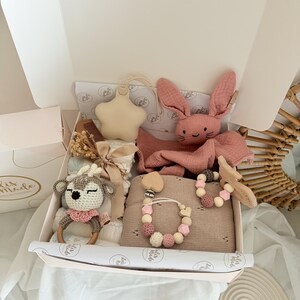 May include: A baby gift box filled with a variety of items. Includes a pink bunny plush, a star-shaped ornament, a crochet deer rattle, a pacifier, and a blanket. The box is lined with tissue paper and has a bow.