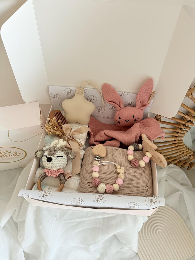 May include: A baby gift box filled with a variety of items. Includes a pink bunny plush, a star-shaped ornament, a crochet deer rattle, a pacifier, and a blanket. The box is lined with tissue paper and has a bow.