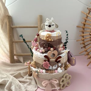 May include: A three-tiered diaper cake decorated with a crocheted deer, pink and brown beads, and a wooden heart. The cake is wrapped in burlap and lace and sits on a beige fabric surface.