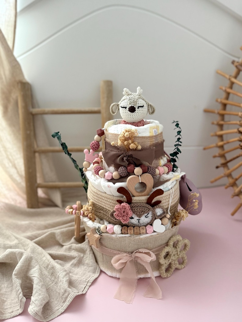 May include: A three-tiered diaper cake decorated with a crocheted deer, pink and brown beads, and a wooden heart. The cake is wrapped in burlap and lace and sits on a beige fabric surface.