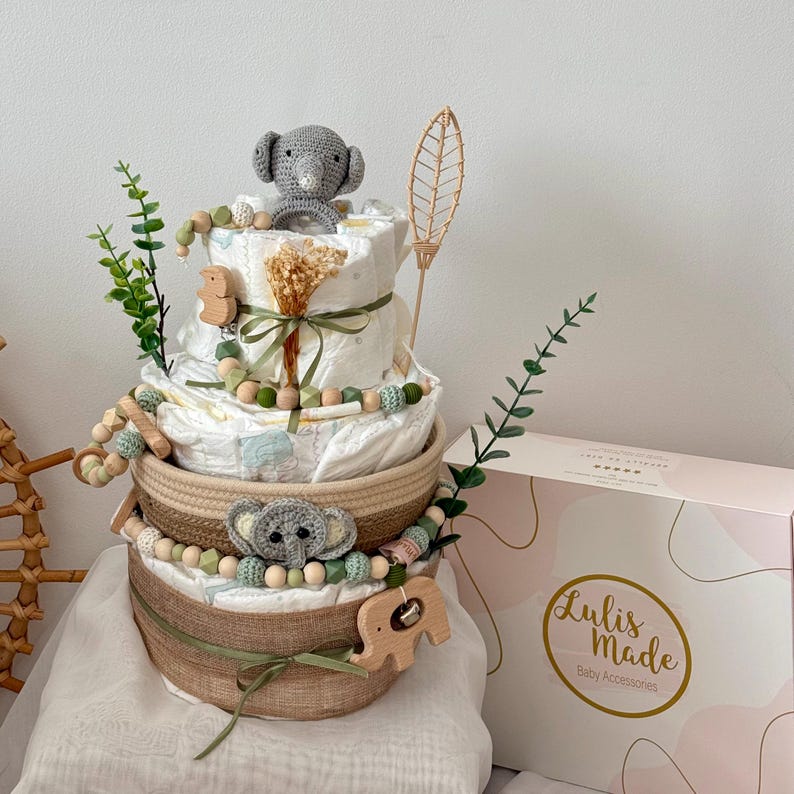 Op de afbeelding: Een drielaags baby shower cadeaupakket met een bosmotief. De mand is versierd met groene bladeren, houten dieren speelgoed en een grijze gehaakte olifant. De mand is bedekt met een houten bladvormige decoratie. De mand staat op een wit oppervlak met een roze doos waarop "Lulis Made Baby Accessories" staat.