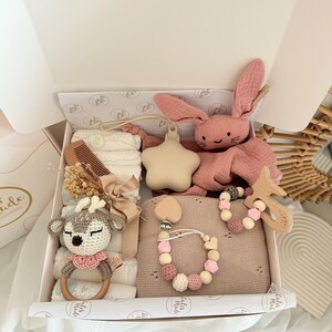 May include: A baby gift box filled with a variety of items. Includes a pink plush bunny, a wooden teether, a pacifier clip, a crochet deer rattle, a comb, and a blanket. The box is white with a decorative interior.