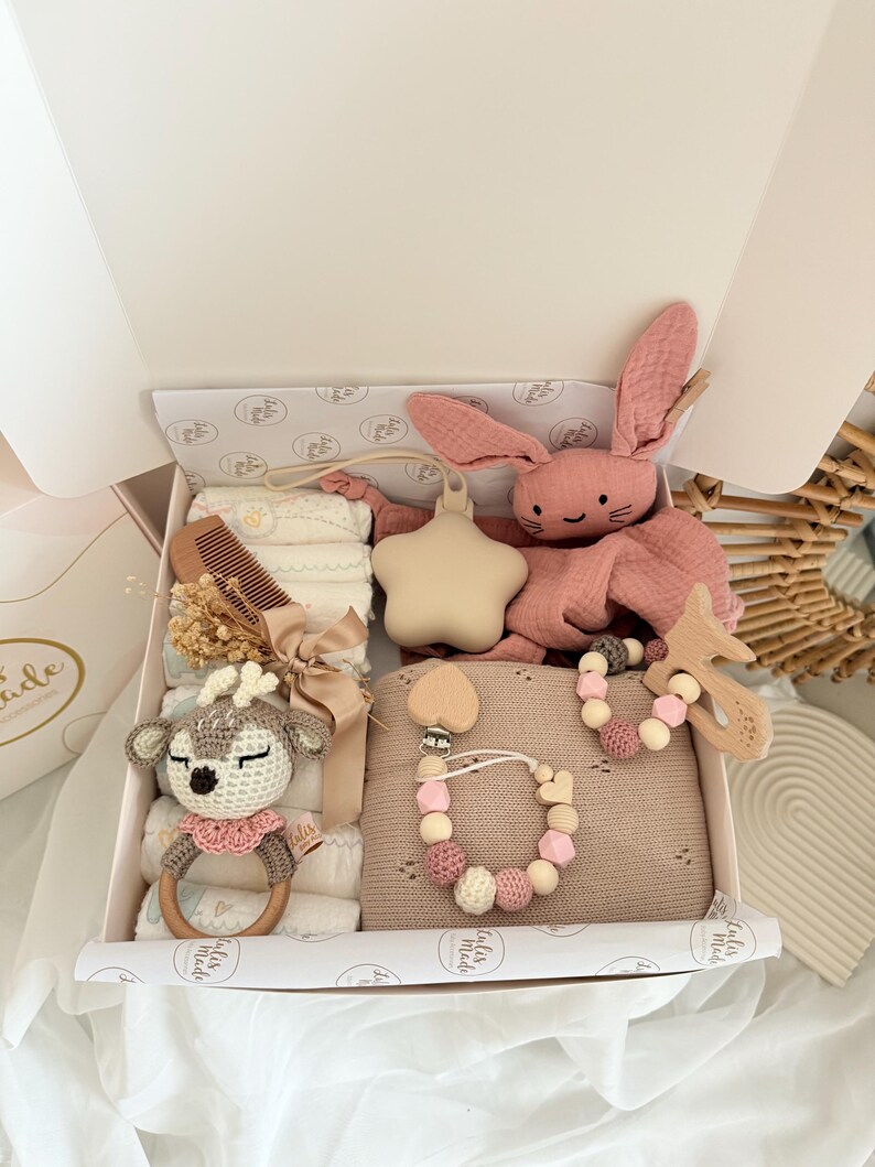 May include: A baby gift box filled with a variety of items. Includes a pink plush bunny, a wooden teether, a pacifier clip, a crochet deer rattle, a comb, and a blanket. The box is white with a decorative interior.