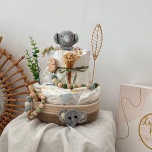 Op de afbeelding: Een baby shower cadeau mand met een neutrale kleurstelling. De mand is gevuld met luiers, een gehaakt olifantenspeelgoed, een houten rammelaar en een groene, bladerrijke tak. De mand staat op een wit oppervlak.