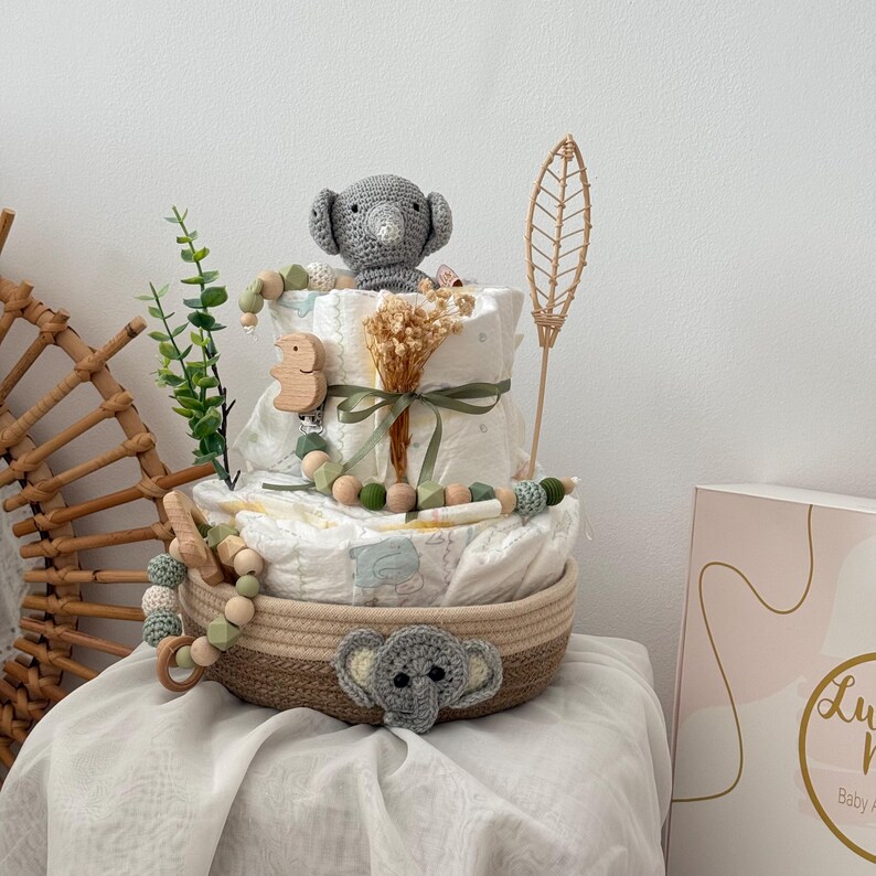 Op de afbeelding: Een baby shower cadeau mand met een neutrale kleurstelling. De mand is gevuld met luiers, een gehaakt olifantenspeelgoed, een houten rammelaar en een groene, bladerrijke tak. De mand staat op een wit oppervlak.